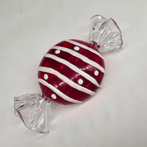 May include: A red and white candy-shaped ornament. The ornament has a round shape with diagonal white stripes and white dots. The ends are clear, translucent, and twisted to resemble a wrapped candy.