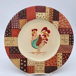 Puede incluir: Plato de cerámica decorativo con una ilustración de gallo. El plato presenta un diseño de patchwork en tonos marrones, naranjas y crema. El texto incluye "Love Begins at Home", "Have a Joyful Day" y "I'm too busy to be organized."