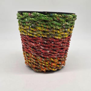 May include: A woven basket with a black rim. The basket is made of colorful, twisted rope in shades of green, yellow, red, and orange. The rope is tightly woven, creating a textured surface. The basket is cylindrical in shape.