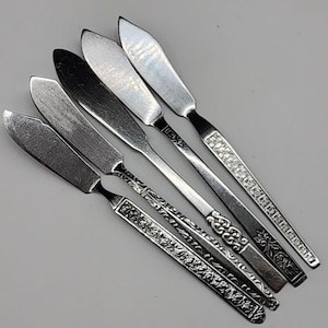 Peut inclure: Six couteaux à beurre argentés vintage avec différents motifs de manche. Les lames sont plates et larges, et les manches présentent des motifs ornés, notamment floraux et géométriques. Les couteaux sont disposés en diagonale sur un fond gris clair.