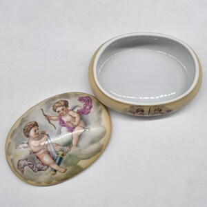May include: An open, oval-shaped trinket box with a lid featuring a painted scene of cherubs. The box is beige with a white interior. The lid's artwork depicts two cherubs with bows and arrows, set against a cloudy background.