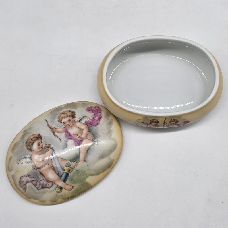 May include: An open, oval-shaped trinket box with a lid featuring a painted scene of cherubs. The box is beige with a white interior. The lid's artwork depicts two cherubs with bows and arrows, set against a cloudy background.