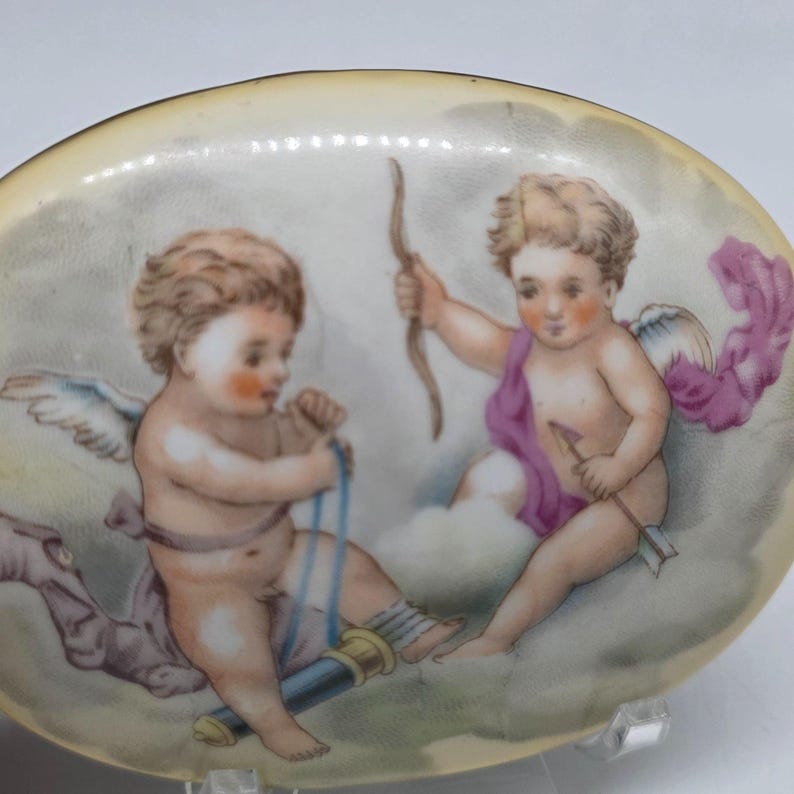 May include: Oval-shaped decorative dish with a painted scene of two cherubs. One cherub holds a bow and arrow, the other holds a telescope. The cherubs are depicted with wings and are set against a cloudy background. The dish has a cream-colored border.