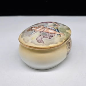 May include: A small, oval-shaped ceramic trinket box with a lid. The lid features a painted scene of a cherub in a cloudscape. The box is primarily cream-colored with a painted design on the side.