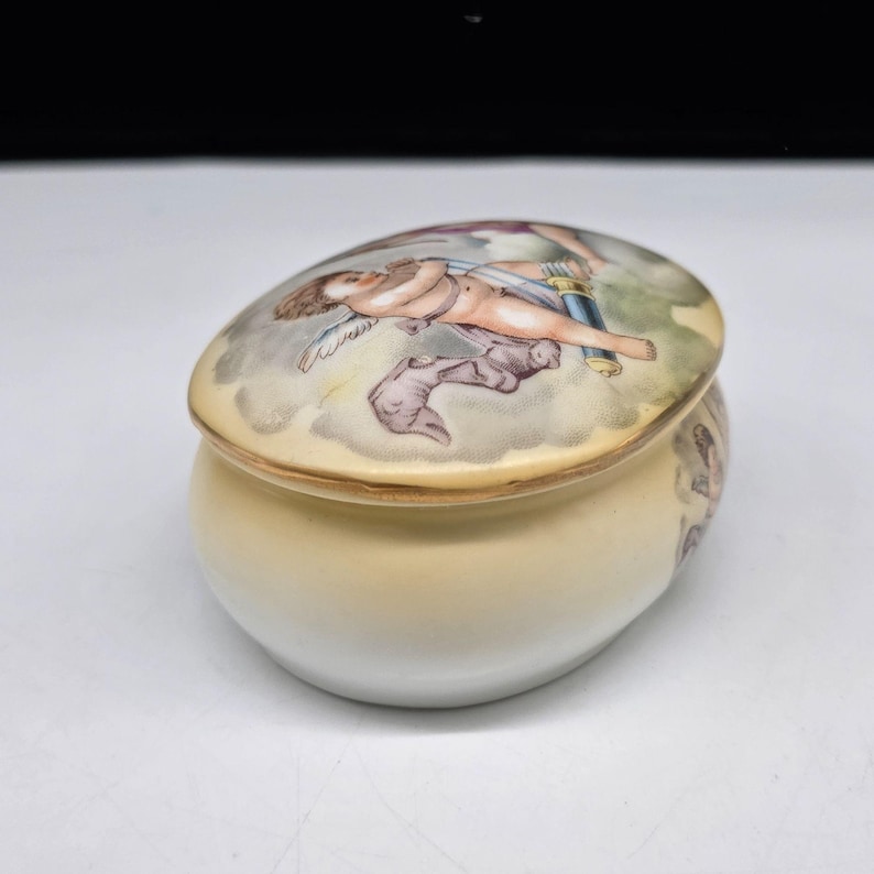 May include: A small, oval-shaped ceramic trinket box with a lid. The lid features a painted scene of a cherub in a cloudscape. The box is primarily cream-colored with a painted design on the side.