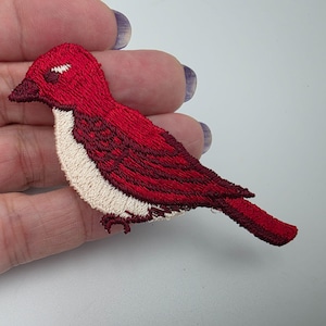 May include: An embroidered patch in the shape of a bird, featuring vibrant red thread for the head, wings, and tail, contrasted with a cream-colored chest and underbelly. The bird has a dark red beak and detailed stitching throughout.