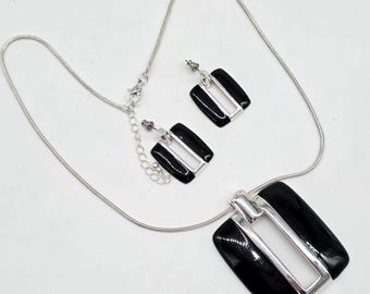 May include: A silver-toned necklace and earring set. The necklace features a large, rectangular pendant with a black enamel finish and a silver-tone frame. The matching earrings have a similar design. The set is displayed on a white background.