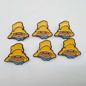 May include: Six identical enamel pins featuring a cartoon character wearing a bright yellow hat and a light blue collar. Each pin has a tan face with simple black outlines for the eyes, nose, and mouth, all enclosed in a dark metal border.