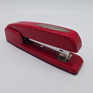 May include: A red stapler with a silver metal staple guide. The stapler is made of a glossy red material and has a curved top. The stapler is designed for office or school use.