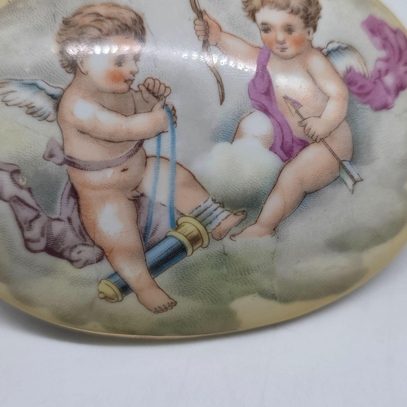 May include: A decorative, oval-shaped porcelain plate featuring a painted scene of two cherubs. One cherub is holding a telescope, while the other holds a bow and arrow. The cherubs are depicted with wings and are set against a cloudy background.
