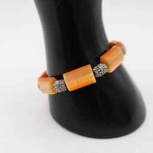 May include: Orange and silver bracelet. The bracelet features rectangular orange beads with silver-colored accents. The bracelet is displayed on a black stand against a white background. The beads have a gradient of orange hues.