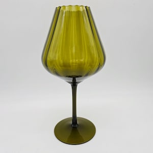 May include: Olive green glass vase with a flared, ribbed top and a long stem. The vase has a rounded base and a dark green stem. The glass has a slight transparency, allowing light to pass through. The vase is a decorative item.