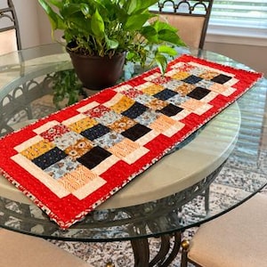 May include: A rectangular quilted table runner featuring a patchwork design of small squares in various patterns and colours, including red, black, yellow, and cream. The runner is bordered by a bright red fabric with a subtle white speckled pattern.