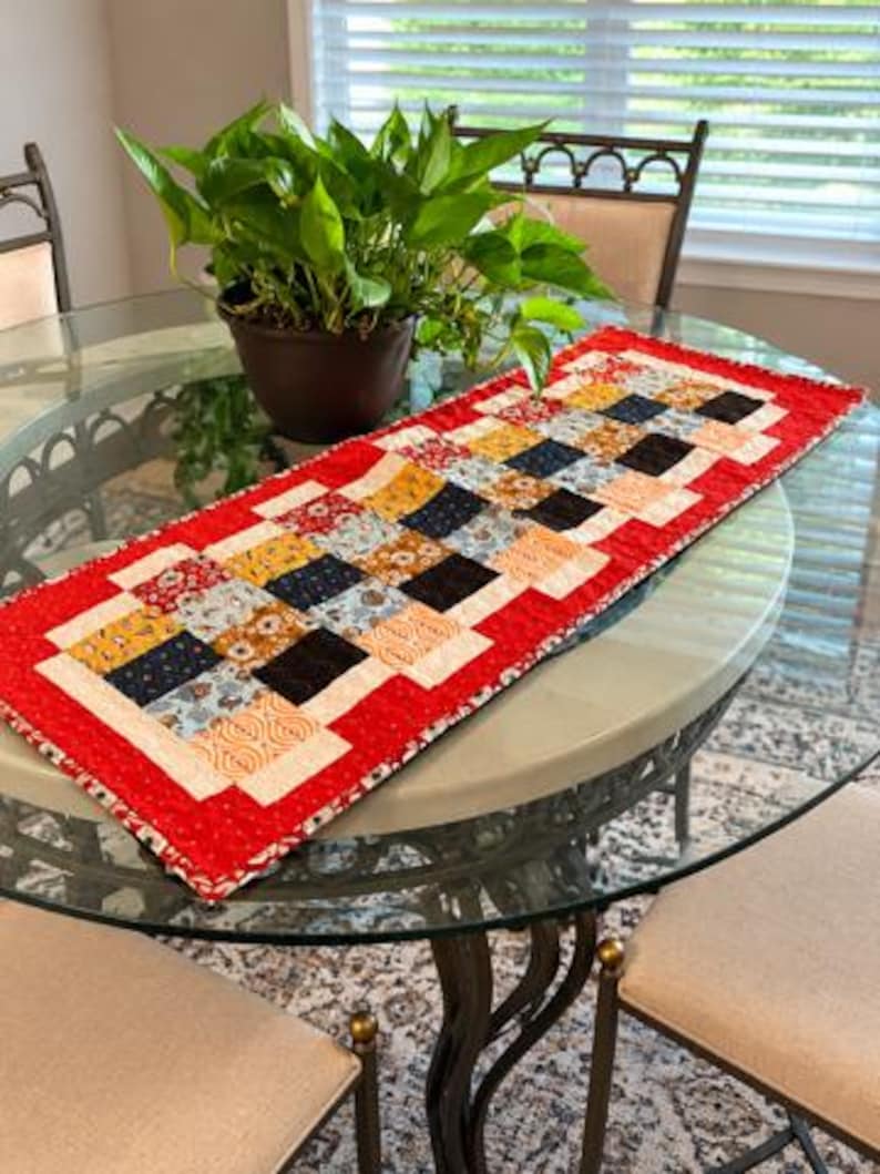 May include: A rectangular quilted table runner featuring a patchwork design of small squares in various patterns and colours, including red, black, yellow, and cream. The runner is bordered by a bright red fabric with a subtle white speckled pattern.