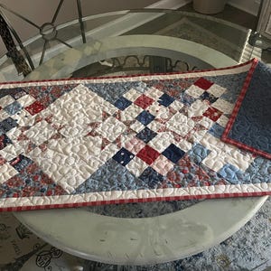 May include: Rectangular quilted table runner with a patchwork design in red, white, and blue. The runner features a repeating pattern of squares and diamonds, with a red and white gingham border. The reverse side is solid blue.