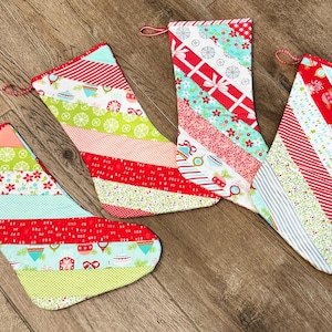 Può includere: Quattro calze di Natale fatte a mano con tessuti a fantasia rossi, verdi e blu. Ogni calza ha un design diverso, con strisce e vari motivi a tema festivo. Sono disposte su una superficie di legno.