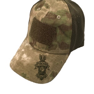 Puede incluir: Una gorra de béisbol de camuflaje A-Tacs con un panel trasero de malla verde. La gorra tiene un parche de velcro en la parte delantera y un logotipo F-Bomb bordado en la visera.