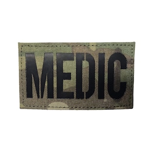 May include: A black and green camouflage fabric patch with the word "MEDIC" printed in black.
