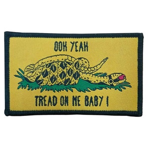 Peut inclure: Un écusson jaune avec une bordure noire représente un serpent à sonnettes enroulé avec une langue rose et le texte "Don't Tread On Me Baby!" en lettres noires.