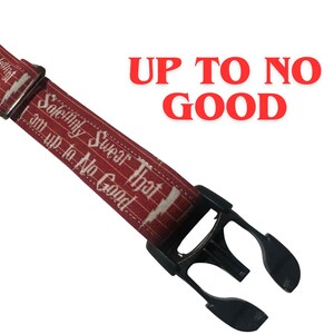 Può includere: Un cordino rosso con testo bianco che recita "Giuro solennemente di non avere buone intenzioni". Il cordino ha un fermaglio in plastica nera e le parole "Up to No Good" in rosso.