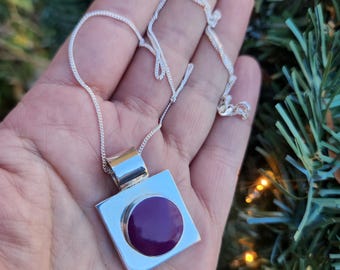 May include: A silver necklace with a square pendant featuring a circular, deep purple center. The pendant is held by a silver chain. The necklace is displayed against a backdrop of green foliage.