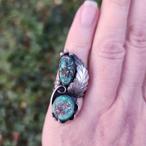 Puede incluir: Anillo de plata con dos piedras turquesas ovaladas con matriz marrón. El anillo tiene un diseño de hoja y pequeñas cuentas de plata. Se lleva en un dedo sobre un fondo verde borroso.