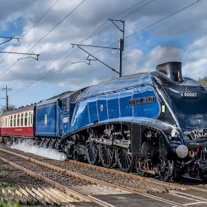 Puede incluir: Una locomotora de vapor azul y negra con el número 60007 en el lateral. El tren se mueve por una vía con un cielo azul nublado en el fondo.