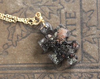 Op de afbeelding: Een gouden ketting met een zwarte en roze puzzelstukje hanger. De hanger is gemaakt van hars en heeft een glinsterende afwerking.