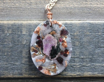 May include: A silver chain necklace with an oval pendant featuring a pink and black crystal encased in clear resin with copper flecks and small red crystals.