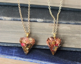 Op de afbeelding: Twee gouden kettingen met een diamantvormige hanger. Elke hanger is gevuld met gouden vlokken, rode en bruine kristallen en andere natuurlijke elementen.