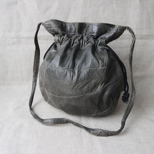 Puede incluir: Un bolso de cuero gris con cordón de ajuste y una correa larga. El bolso tiene un diseño acolchado texturizado.