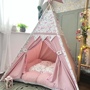 May include: A pink and white floral teepee tent with a matching floor cushion. The tent has a decorative banner and pom-pom trim. The tent is set up indoors on a gray rug.