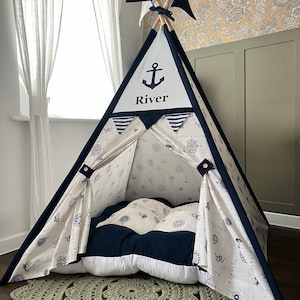 May include: A navy and white teepee tent with an anchor graphic and the name "River". The tent has a nautical theme with compass and map patterns. Includes a matching navy and white cushion and a green crocheted rug.