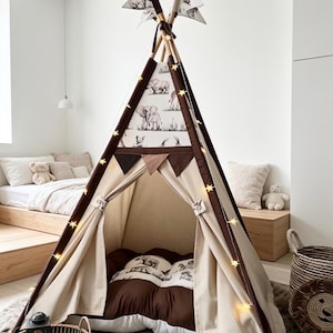 May include: A beige and brown teepee tent with a decorative elephant pattern. The tent has a brown trim, star-shaped lights, and decorative flags at the top. Inside, there are pillows and a brown blanket. The tent is set up in a child's bedroom.
