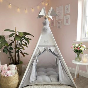 May include: A white and gray teepee tent with the name "Sophia" printed on the front. The tent has gray flags and pom-poms on top, and gray fabric accents. A woven basket with stuffed animals sits near the tent.