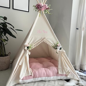 May include: A cream-colored teepee tent with a pink and white padded floor. The tent is decorated with floral arrangements and the name "Ela Nur" in pink script. The tent is set up indoors on a patterned rug.