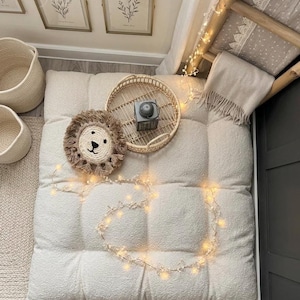 May include: A cozy, neutral-toned interior scene featuring a large, tufted floor cushion. A round, woven tray holds a small gray object. A lion-shaped decorative pillow and a string of fairy lights add charm. Woven baskets and framed botanical prints complete the space.