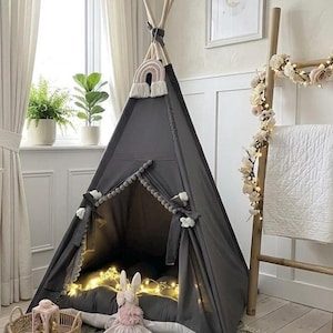 May include: A gray teepee tent with a rainbow decoration and string lights inside. The tent is set up in a room with a window, plants, and a wooden ladder decorated with flowers. A stuffed bunny and a woven basket are in front of the tent.