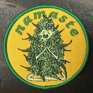 May include: A green and yellow embroidered patch with the word "Namaste" written around the edge. In the center of the patch is a cartoon figure meditating, surrounded by green marijuana leaves.
