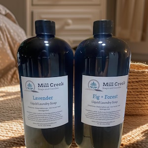 May include: Two dark blue bottles of Mill Creek Liquid Laundry Soap. One is labeled "Lavender" and the other "Fig + Forest." Each bottle has a black cap and a white label with product information. The bottles are sitting on a woven surface.