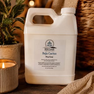 May include: A white plastic jug of Mill Creek Soaps and Candles Baja Cactus Mop Soap. The label features a logo, product name, directions to mix 4 ounces with 1 gallon of water, ingredients, website, and a net weight of 32 ounces (908g).