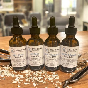 May include: Four amber glass bottles of beard oil with black droppers. Each bottle has a white label with the text "Mill Creek Soaps and Candles" and a different scent name: "Ash + Stone", "Lost Sailor", "Santal + Cardamom", and "Smoke & Society".