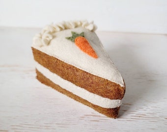 May include: A felt carrot cake slice with white frosting and a green carrot on top. The cake is on a white wooden surface.