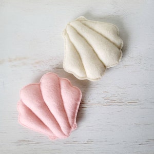 May include: Two felt seashells, one pink and one white, on a white wooden background.