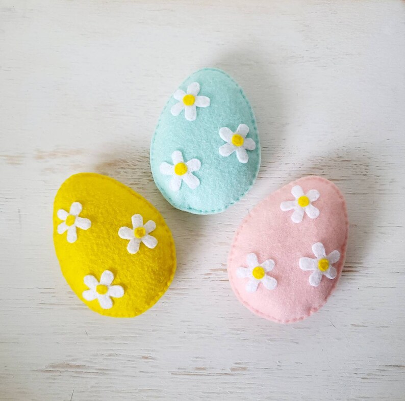May include: Three felt Easter eggs decorated with white daisies. The eggs are yellow, light blue, and pink.