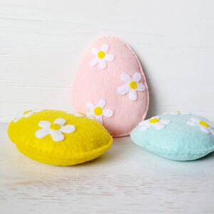 May include: Three felt Easter eggs, one pink, one yellow, and one light blue, each decorated with white felt daisies.