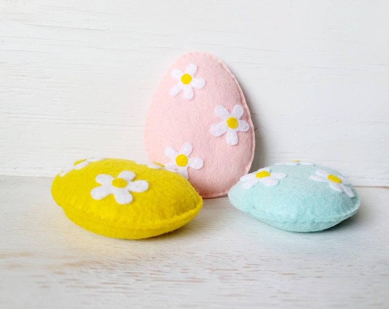 May include: Three felt Easter eggs, one pink, one yellow, and one light blue, each decorated with white felt daisies.