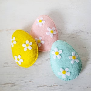May include: Three felt Easter eggs decorated with white felt flowers. The eggs are pink, yellow, and light blue.