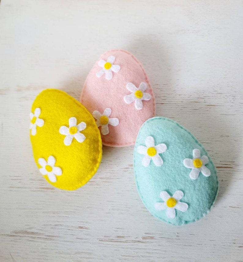 May include: Three felt Easter eggs decorated with white felt flowers. The eggs are pink, yellow, and light blue.