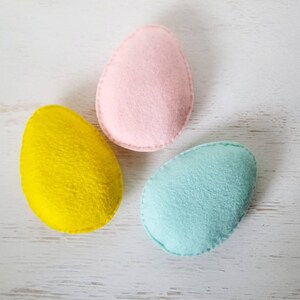 May include: Three felt Easter eggs in pink, yellow, and light blue. The eggs are arranged in a triangle shape on a white wooden surface.